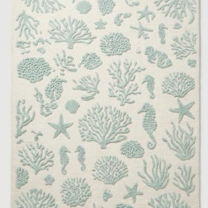 Puede incluir: Una alfombra rectangular con fondo color crema y un patr&oacute;n repetitivo de motivos marinos en relieve de color azul verdoso claro, que incluye diversas formaciones de coral, estrellas de mar, caballitos de mar y conchas distribuidas.