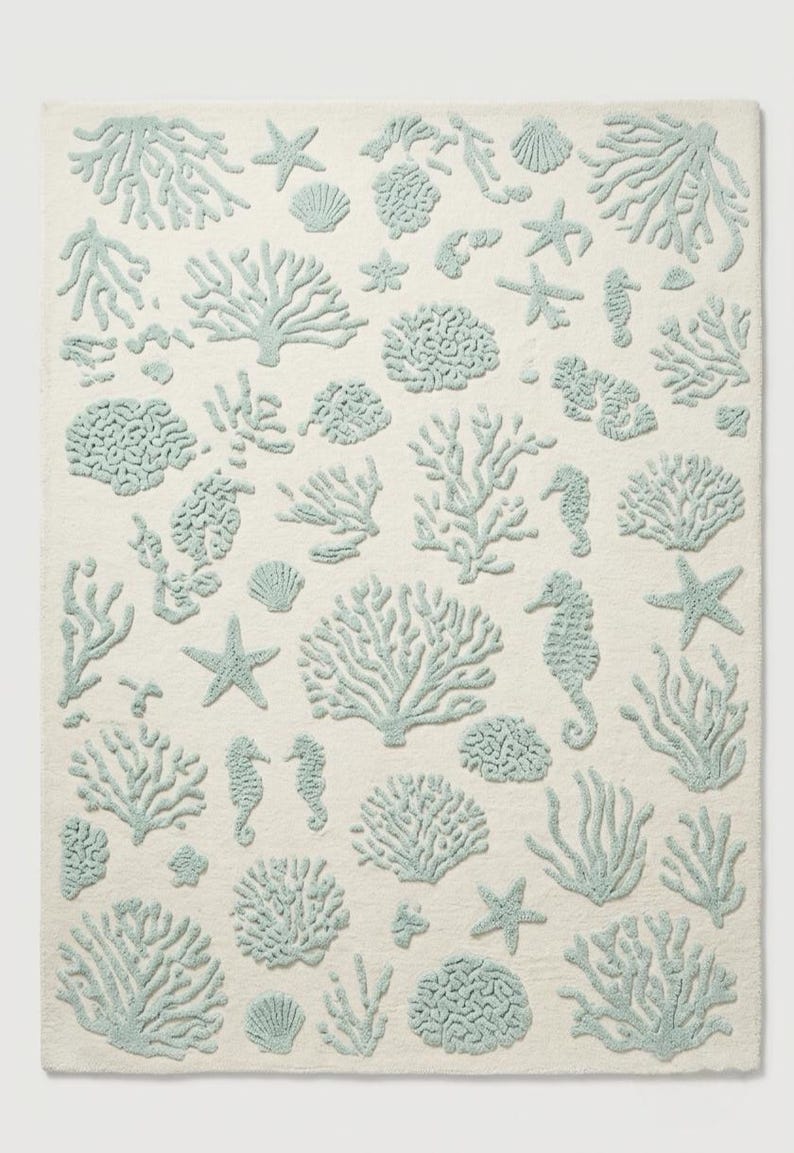Puede incluir: Una alfombra rectangular con fondo color crema y un patr&oacute;n repetitivo de motivos marinos en relieve de color azul verdoso claro, que incluye diversas formaciones de coral, estrellas de mar, caballitos de mar y conchas distribuidas.