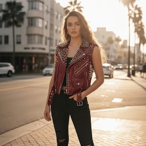 May include: A burgundy sleeveless leather vest with silver metal studs and zips. The vest has a classic biker jacket design, with a notched collar and a fitted silhouette. The model is wearing black jeans.