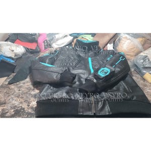 Puede incluir: Una chaqueta negra con detalles en color cian brillante, cuello alto y puños negros acanalados. Las mangas presentan un parche cuadrado con un gráfico de circuito impreso en cian y la letra N. Una franja cian vertical recorre la cremallera.