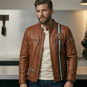 Puede incluir: Una chaqueta de cuero marrón con cuello mao, cierre de cremallera frontal y una franja vertical azul y blanca en el lado izquierdo. La chaqueta incluye un parche de Ford Performance, un parche de bandera a cuadros y bolsillos con cremallera.