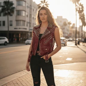 May include: A burgundy sleeveless leather vest with silver metal studs and zips. The vest has a classic biker jacket design with a notched collar and a belt at the waist. The model is wearing black jeans.