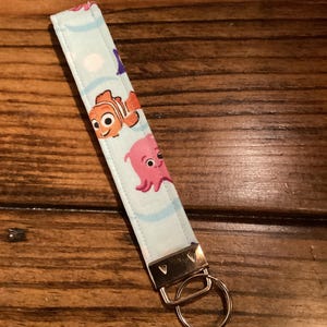 May include: A blue and white fabric keychain with a cartoon image of Nemo and a pink octopus. The keychain has a gold metal ring.