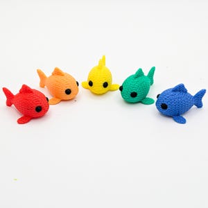 May include: A set of five small, textured fish figurines in red, orange, yellow, green, and blue. Each fish features a distinct cross-hatched scale pattern, prominent fins, and two round, black eyes, arranged in a semi-circle on a plain surface.