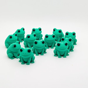 May include: A collection of small, teal-colored frog figurines with black eyes. The frogs are arranged on a white surface. The figurines have a textured, knitted appearance, and are all facing forward.