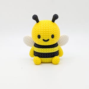May include: A yellow and black bee figurine with a smiling face, black antennae, and white wings. The bee has a textured, knitted appearance. The figurine is sitting on a white surface.