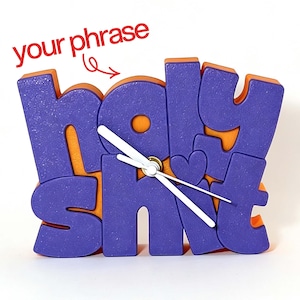 Può includere: Un orologio da scrivania viola e arancione con il testo "holy shit" in un carattere grassetto e arrotondato, con un piccolo cuore integrato nel design. L'orologio ha tre lancette bianche centrate sul quadrante e il testo "your phrase" sopra.