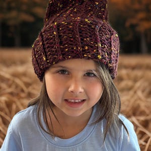 Puede incluir: Un gorro de punto color granate oscuro con una textura acanalada gruesa y pequeñas motas coloridas de hilo amarillo, naranja y azul tejidas en la tela. El gorro tiene un borde doblado y una corona redondeada, hecho de hilo grueso.