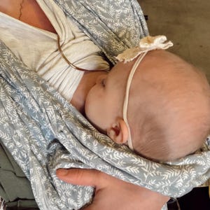 May include: A baby carrier wrap featuring a light blue fabric with a white botanical leaf pattern. The wrap is secured with a metallic ring. A baby is positioned inside the carrier, wearing a thin headband with a small, cream-colored fabric flower.