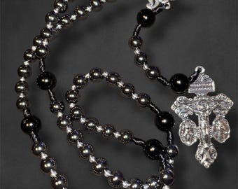 May include: A rosary with dark gray and black beads. The beads are strung together with black and white string. A silver-colored crucifix and a decorative piece with the letter "M" are attached.