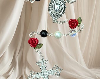 May include: A rosary with a silver cross pendant adorned with clear crystals. The rosary features black, white, and iridescent beads, along with red rose accents. The design includes a decorative heart-shaped centerpiece.