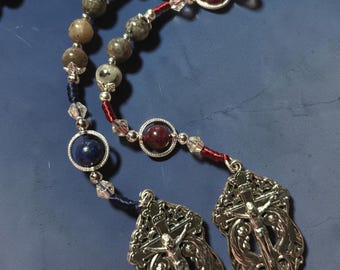 May include: A rosary featuring grey stone beads, clear crystal accents, and silver-toned metal components. The design includes two ornate silver crucifixes, two heart-shaped medals, and circular frames containing deep blue and dark red spherical beads.