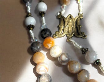 May include: A rosary featuring multi-colored stone beads in shades of white, gray, orange, and tan, strung on white cord. The piece includes small clear accent beads, antique brass-toned metal spacers, and a central brass-toned medal depicting a figure.