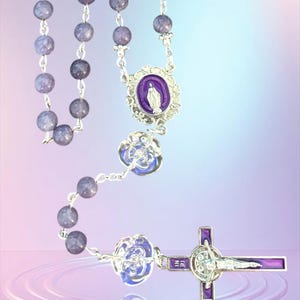 May include: A rosary with lavender-colored beads and silver-toned metal accents. The rosary features a purple and silver cross and a pendant with a depiction of the Virgin Mary. The beads are a marbled lavender color.