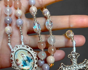 May include: A rosary with iridescent beads in shades of pink, lavender, and white. The rosary features a silver-toned cross and a decorative oval pendant with a religious image. The beads are strung on a white cord.