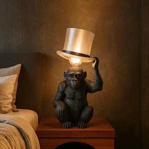 Puede incluir: Una lámpara de mesa con forma de mono negro con una pantalla de sombrero de copa. La lámpara presenta una escultura detallada de un mono sosteniendo el sombrero sobre su cabeza, con una bombilla que se ilumina desde adentro. La lámpara está colocada sobre una mesita de noche de madera.