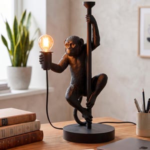 Pu&ograve; includere: Una lampada da tavolo a forma di scimmia nera con una lampadina. La lampada tiene un palo e la lampadina &egrave; nella sua mano. La lampada &egrave; su una scrivania di legno con un laptop, libri e un taccuino.