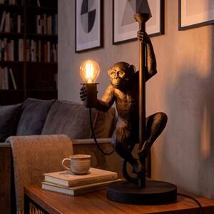 Pu&ograve; includere: Lampada da tavolo decorativa a forma di scimmia che tiene una lampadina. La lampada &egrave; color bronzo e si trova su un tavolo di legno. La scena comprende libri, una tazza e opere d'arte incorniciate sullo sfondo.