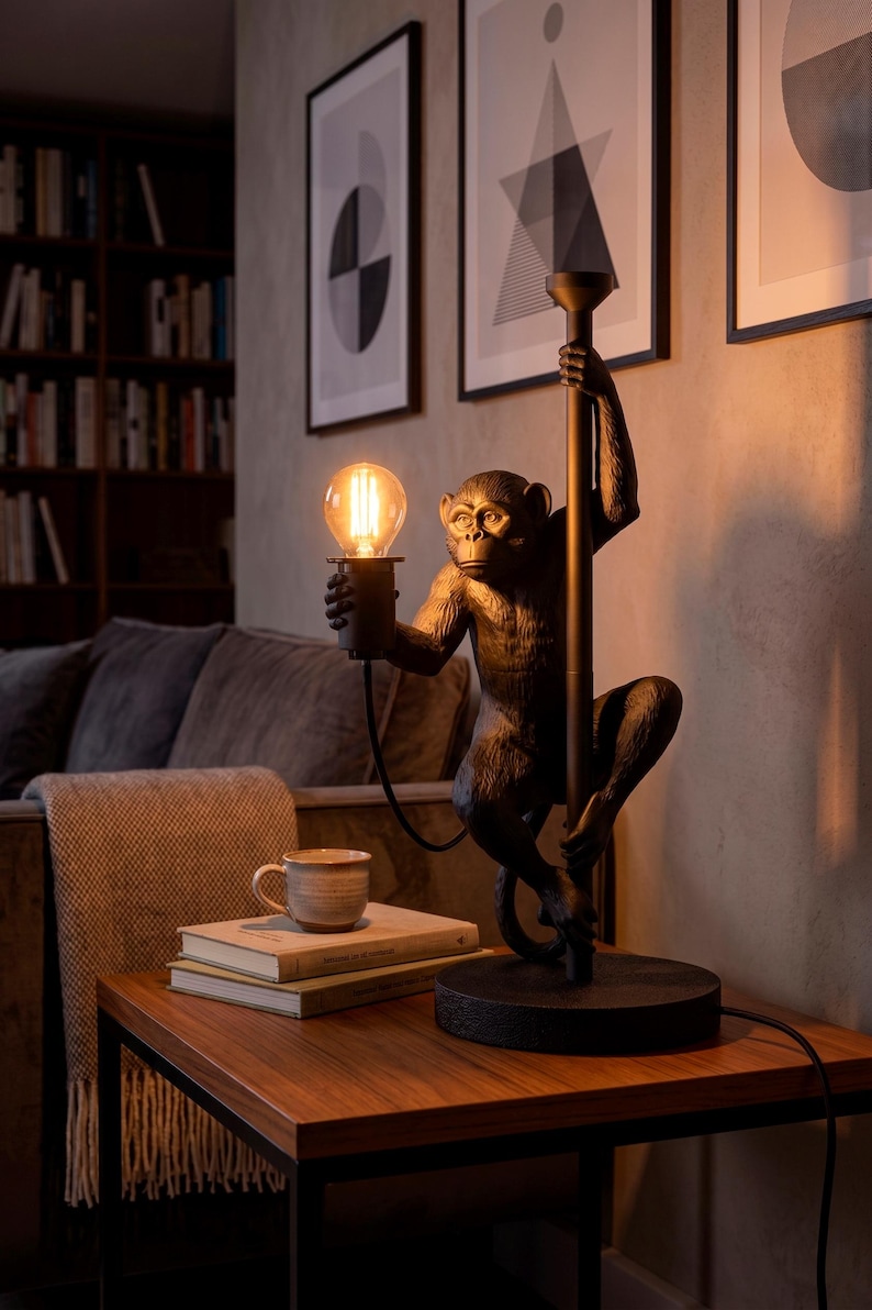 Pu&ograve; includere: Lampada da tavolo decorativa a forma di scimmia che tiene una lampadina. La lampada &egrave; color bronzo e si trova su un tavolo di legno. La scena comprende libri, una tazza e opere d'arte incorniciate sullo sfondo.