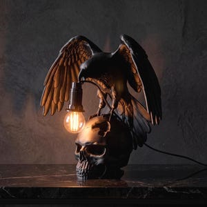 Puede incluir: Una lámpara de mesa con un águila negra de alas extendidas posada sobre un cráneo humano. El águila sostiene en su pico un casquillo plateado que contiene una bombilla redonda de estilo Edison que ilumina la oscura escultura.