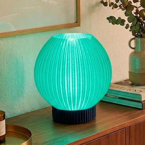 Puede incluir: Una lámpara de mesa esférica de color turquesa con detalles acanalados verticales. La lámpara se asienta sobre una base cilíndrica azul oscuro. La lámpara está iluminada, emitiendo un brillo suave. La lámpara está sobre una superficie de madera, con un cuadro enmarcado y un jarrón en el fondo.