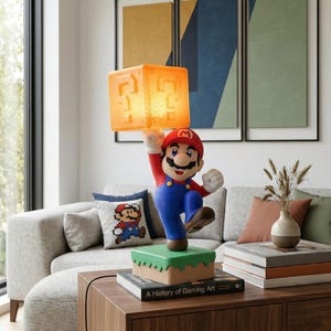 May include: A Mario-themed lamp featuring the character holding a glowing orange question mark block. The lamp is on a green and brown base, with a book titled "A History of Gaming Art" nearby. The lamp is a decorative item.