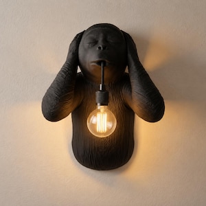 Könnte beinhalten: Eine mattschwarze Wandleuchte in Form eines Gorillakopfes und -oberkörpers. Die Figur hält ihre Hände über die Ohren und hat die Augen geschlossen, während eine leuchtende Edison-Glühbirne an einem schwarzen Kabel aus ihrem Mund hängt.