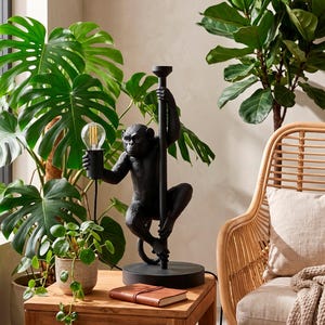 Pu&ograve; includere: Lampada da tavolo nera a forma di scimmia che tiene una lampadina. La lampada &egrave; su una base rotonda e ha un palo. La lampada &egrave; su un tavolo di legno con un taccuino rilegato in pelle. Lo sfondo comprende piante e una sedia in vimini.