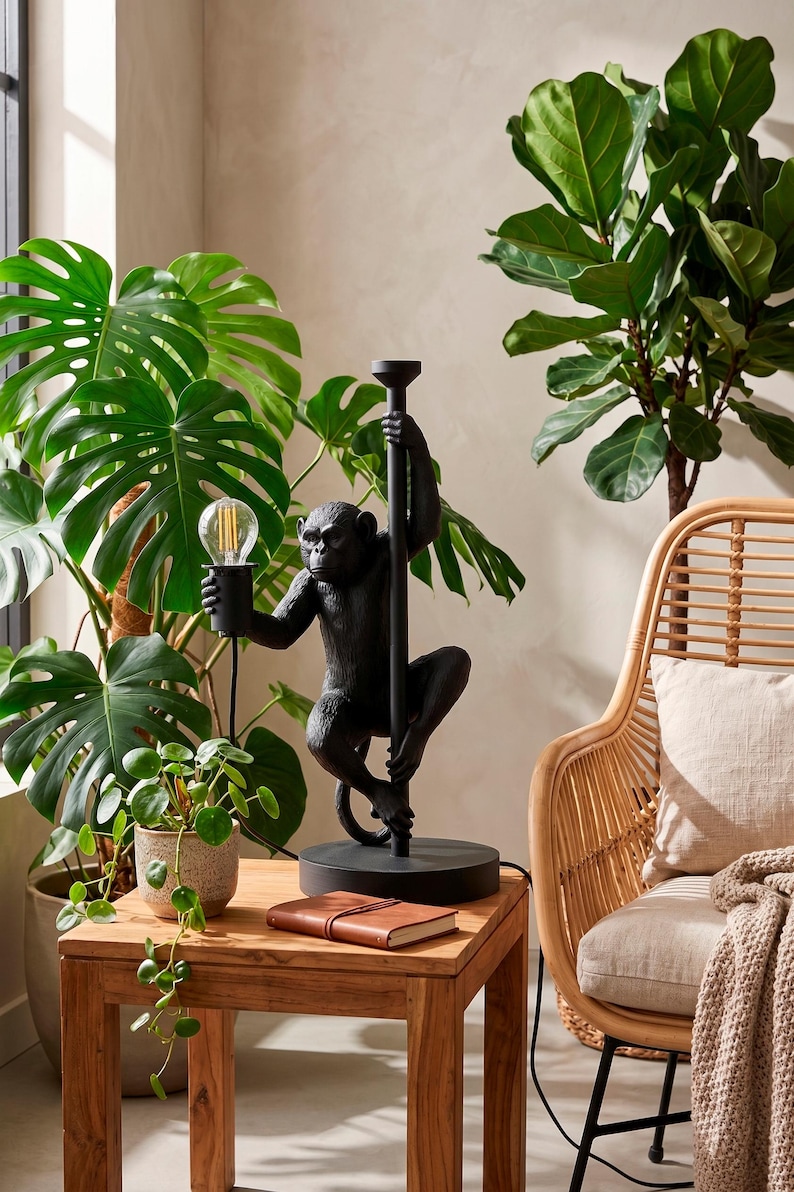 Pu&ograve; includere: Lampada da tavolo nera a forma di scimmia che tiene una lampadina. La lampada &egrave; su una base rotonda e ha un palo. La lampada &egrave; su un tavolo di legno con un taccuino rilegato in pelle. Lo sfondo comprende piante e una sedia in vimini.