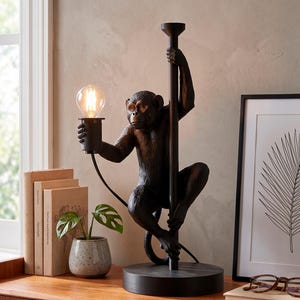Pu&ograve; includere: Una lampada da tavolo a forma di scimmia nera con una lampadina. La scimmia tiene un palo e la lampadina. La lampada &egrave; su una base rotonda. La lampada &egrave; su una superficie di legno con libri e una pianta.