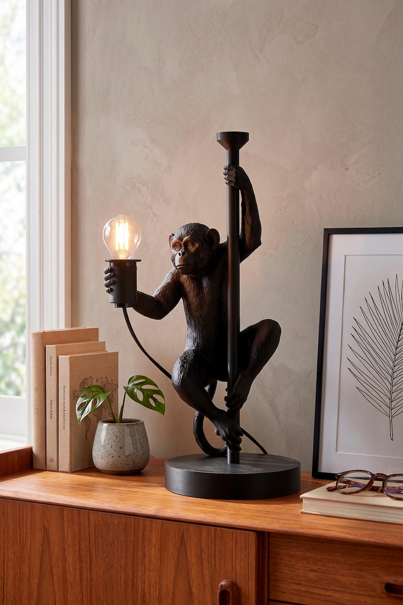 Pu&ograve; includere: Una lampada da tavolo a forma di scimmia nera con una lampadina. La scimmia tiene un palo e la lampadina. La lampada &egrave; su una base rotonda. La lampada &egrave; su una superficie di legno con libri e una pianta.