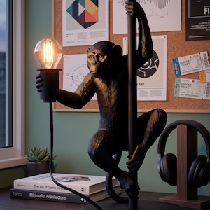 Pu&ograve; includere: Una lampada da scrivania nera a forma di scimmia con una lampadina in stile vintage. La scimmia tiene un palo con una mano e la lampadina con l'altra. La lampada &egrave; su una scrivania con libri e una pianta succulenta.