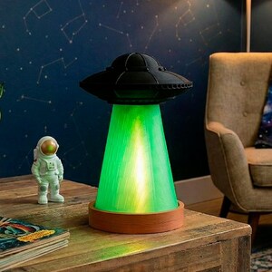 May include: A black UFO-shaped lamp emitting a bright green light beam. The lamp is on a brown base, placed on a wooden table. A small astronaut figurine and a vintage book are also on the table. The background is a dark blue wall with white stars.