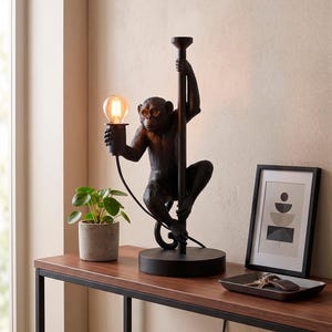 Pu&ograve; includere: Lampada da tavolo nera a forma di scimmia che tiene una lampadina. La lampada &egrave; su un tavolo marrone scuro con una piccola pianta in vaso e una stampa artistica astratta incorniciata. La base della lampada &egrave; rotonda e la scimmia si arrampica su un palo.