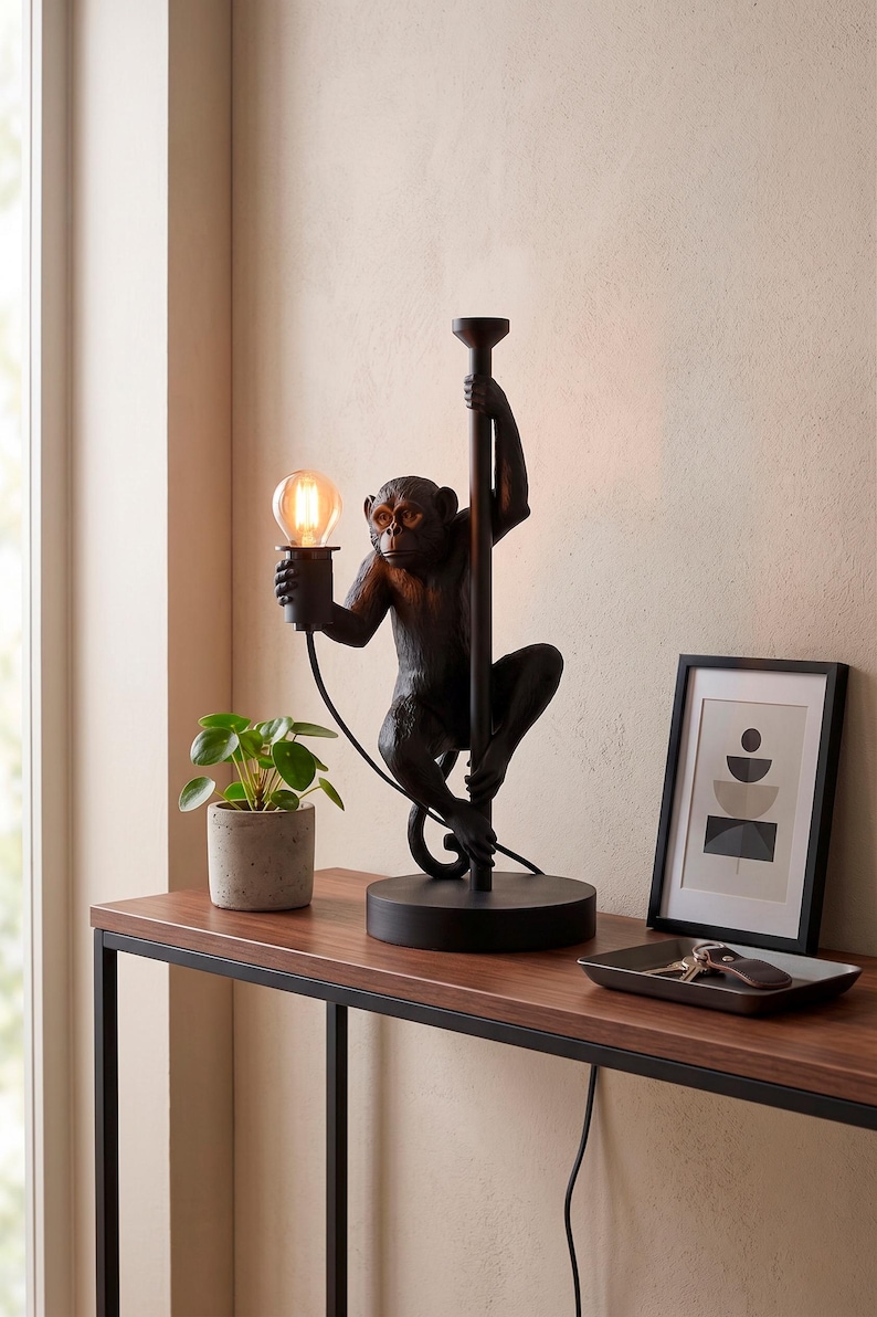 Pu&ograve; includere: Lampada da tavolo nera a forma di scimmia che tiene una lampadina. La lampada &egrave; su un tavolo marrone scuro con una piccola pianta in vaso e una stampa artistica astratta incorniciata. La base della lampada &egrave; rotonda e la scimmia si arrampica su un palo.