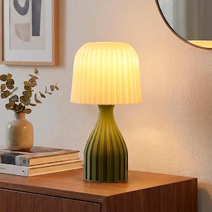 May include: A green and cream-colored table lamp with a pleated lampshade. The lamp has a ribbed, vase-shaped base. The lamp is on a wooden surface, with a vase of greenery and books in the background.