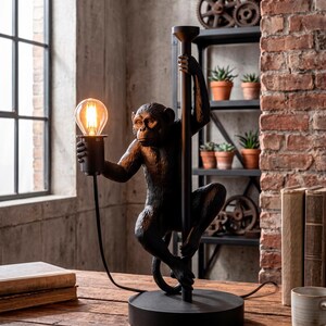 Pu&ograve; includere: Una lampada da tavolo nera a forma di scimmia con una lampadina in stile vintage. La figura della scimmia si aggrappa a un palo nero. La lampada poggia su una base rotonda nera. Lo sfondo include un muro di mattoni e una finestra.