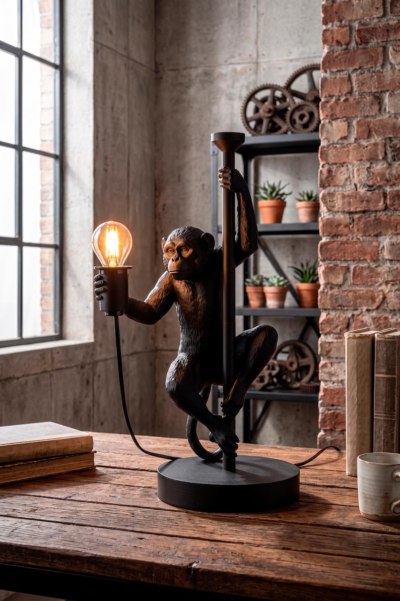 Pu&ograve; includere: Una lampada da tavolo nera a forma di scimmia con una lampadina in stile vintage. La figura della scimmia si aggrappa a un palo nero. La lampada poggia su una base rotonda nera. Lo sfondo include un muro di mattoni e una finestra.