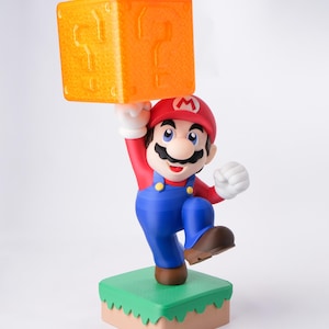 May include: A figurine of Mario, a character from the Super Mario video game series, holding an orange question mark block above his head. Mario is wearing a red hat, red shirt, blue overalls, and brown shoes. He stands on a green and tan base.