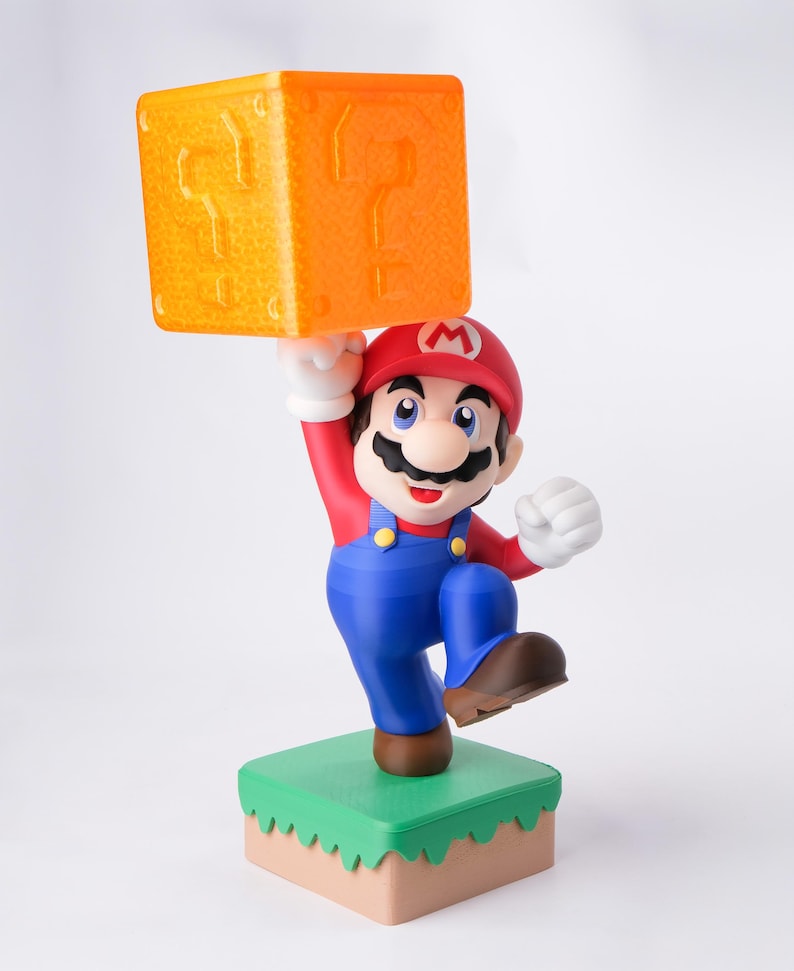 May include: A figurine of Mario, a character from the Super Mario video game series, holding an orange question mark block above his head. Mario is wearing a red hat, red shirt, blue overalls, and brown shoes. He stands on a green and tan base.
