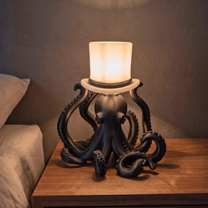 Op de afbeelding: Een zwarte octopuslamp met een witte lampenkap in de vorm van een hoge hoed. De lamp staat op een houten nachtkastje. De tentakels van de octopus krullen rond de basis van de hoed. Een decoratieve lamp.