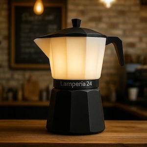 May include: A black and white coffee pot-shaped lamp with the text "Lamperia 24" on the base. The top section glows with a warm light. The lamp is on a wooden surface, with a blurred background of a cafe.
