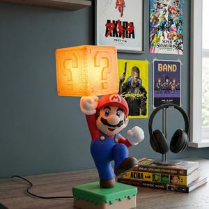 May include: A Mario-themed lamp featuring the iconic character holding a glowing question mark block. The lamp has a red hat, blue overalls, and a green base. The background includes video game posters and shelves with collectibles.