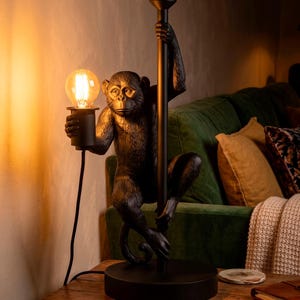 Pu&ograve; includere: Lampada da tavolo nera a forma di scimmia con una lampadina. La scimmia tiene un palo nero con una mano e la lampadina con l'altra. La lampada &egrave; su un tavolo di legno.