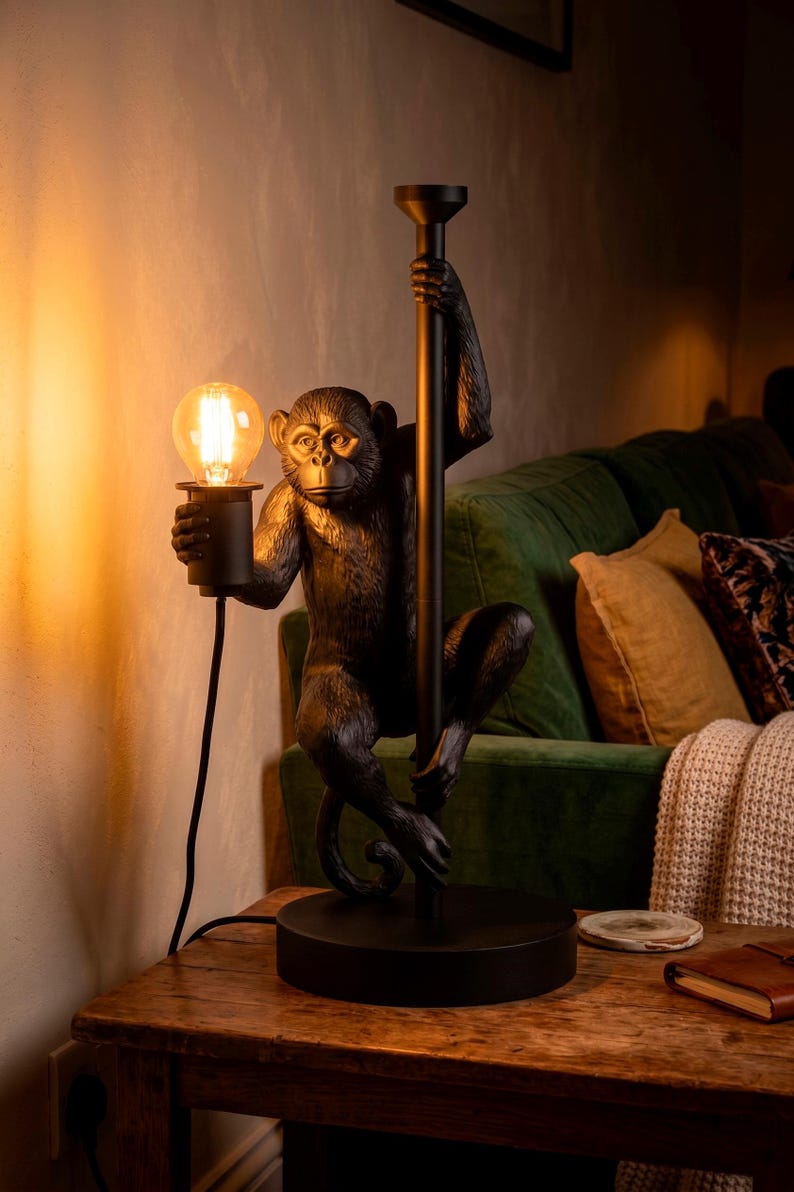 Pu&ograve; includere: Lampada da tavolo nera a forma di scimmia con una lampadina. La scimmia tiene un palo nero con una mano e la lampadina con l'altra. La lampada &egrave; su un tavolo di legno.