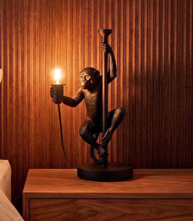 Pu&ograve; includere: Lampada da tavolo decorativa a forma di scimmia, rifinita in bronzo. La scimmia tiene in mano una lampadina e un palo. La lampada &egrave; su una base rotonda, posta su un comodino in legno. Lo sfondo &egrave; una parete in legno.