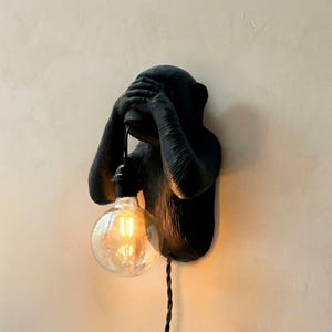 Puede incluir: Aplique de pared negro con forma de mono con las manos cubriendo los ojos. La lámpara tiene una bombilla redonda y transparente que emite un brillo cálido. El aplique está montado en una pared de color neutro y el cable eléctrico es visible.
