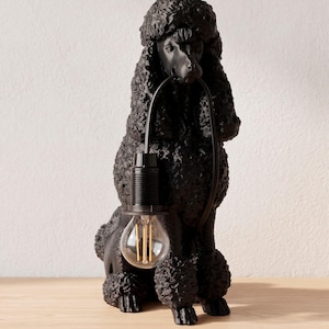 May include: Black poodle-shaped lamp with a curly coat and a light bulb held in its mouth. The lamp has a black cord and a standard light bulb socket. The lamp is sitting on a wooden surface.