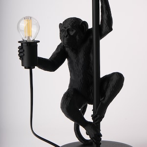 Pu&ograve; includere: Lampada da tavolo nera a forma di scimmia. La lampada presenta una figura di scimmia che tiene un palo nero con una lampadina. La lampada ha una base rotonda e un cavo nero. La lampadina &egrave; trasparente con un filamento visibile.