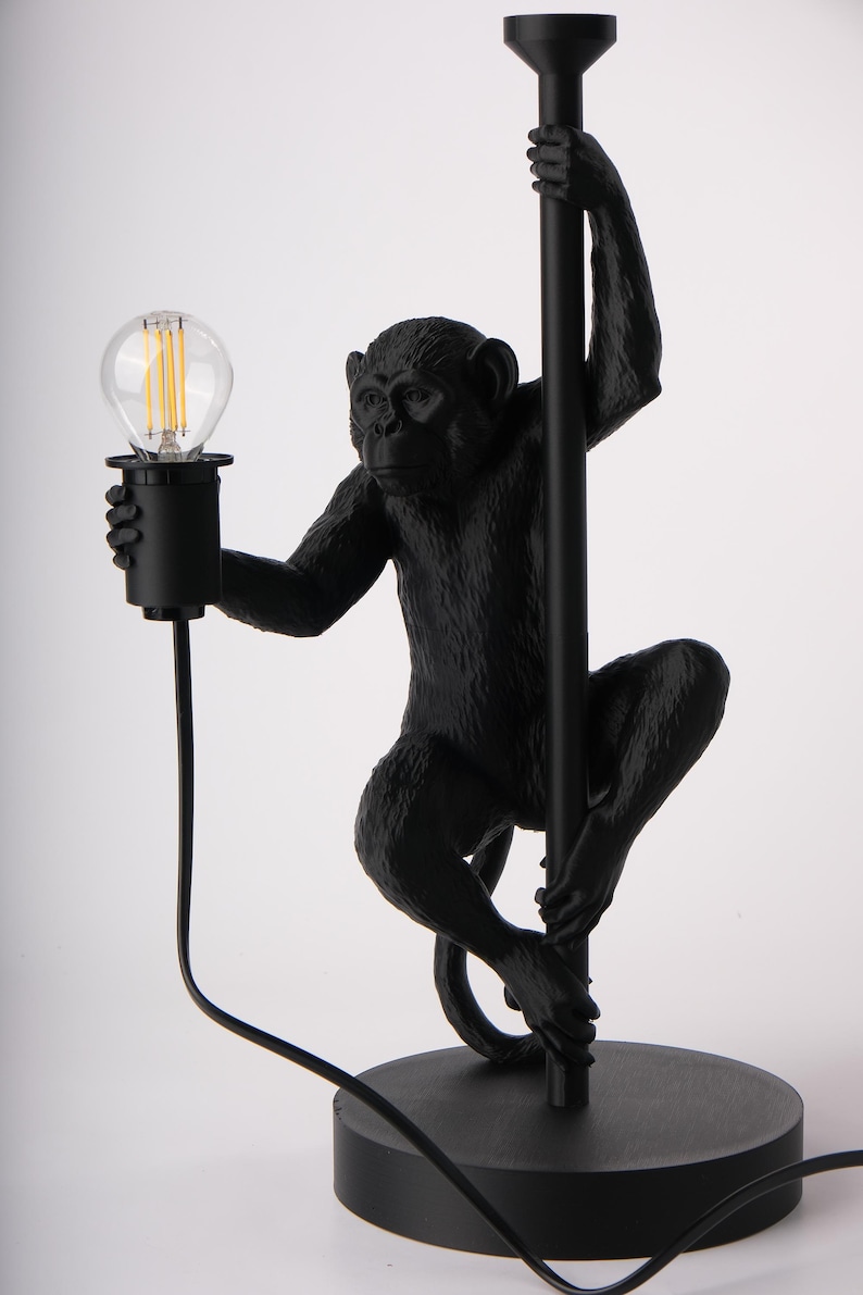 Pu&ograve; includere: Lampada da tavolo nera a forma di scimmia. La lampada presenta una figura di scimmia che tiene un palo nero con una lampadina. La lampada ha una base rotonda e un cavo nero. La lampadina &egrave; trasparente con un filamento visibile.